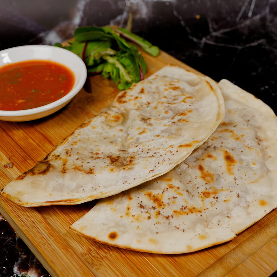 Two grilled flatbreads with a side of red sauce and greens on a wooden board.