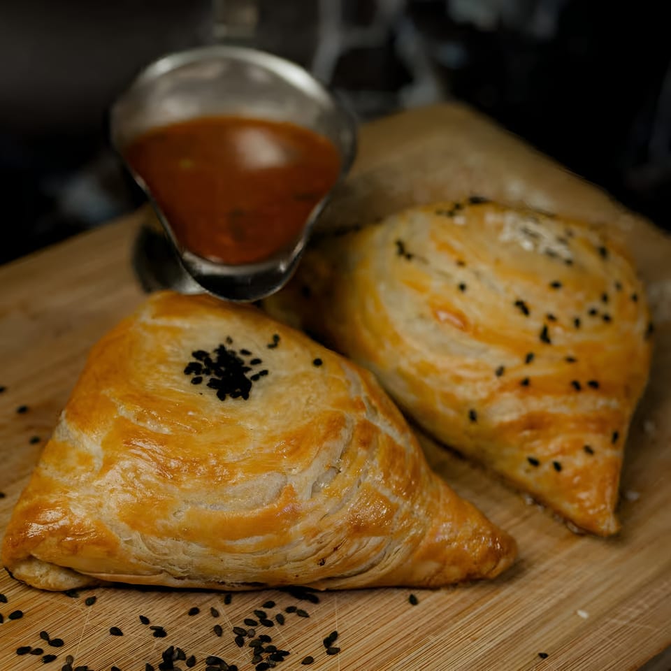 Two golden-brown samosas with black sesame seeds on a wooden board, with a gravy boat.
